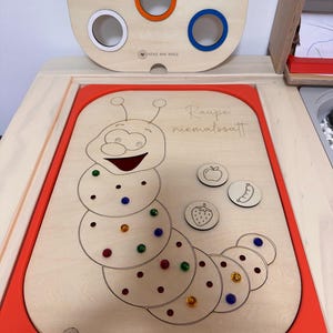 Könnte beinhalten: Holz-Lernspielzeug mit einem Raupen-Design, farbigen Stiften und fruchtförmigen Teilen. Der Text "Raupe niemals satt" ist sichtbar. Das Spielzeug enthält einen oberen Bereich mit farbigen Löchern zum Zuordnen.