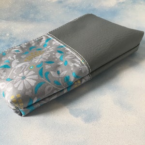 May include: A gray and white fabric pouch with a floral pattern and a gray leather flap. The pouch is closed and lying on a white surface.