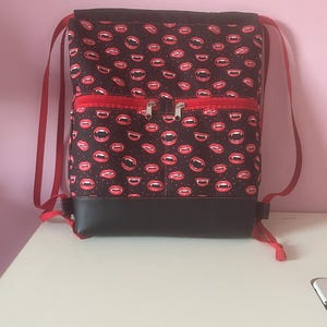 May include: A black backpack with a red zipper pocket and red straps. The backpack is patterned with red lips and fangs on a black background. The bottom of the backpack is black.