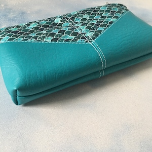 May include: Teal zippered pouch with a mermaid scale pattern on the front. The pouch is made of faux leather and has a white stitching detail.