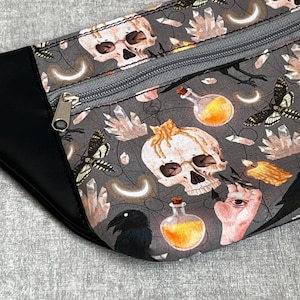 May include: A gray fanny pack with a black leather trim and a silver zipper. The fabric features a repeating pattern of skulls, butterflies, crystals, potion bottles, and a raven. The design has a spooky, gothic theme.