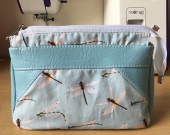 May include: A light blue zippered pouch with a flap closure. The pouch is decorated with a dragonfly print and has a white zipper. The pouch is sitting on a wooden surface. The text "Fabric is cheaper than therapy" is visible on a sticker.