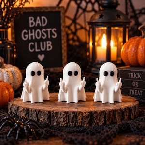 May include: Three white ghost figurines with black eyes, each holding up both middle fingers. The ghosts are arranged in a row on a circular wooden slice. The background features a sign that reads BAD GHOSTS CLUB with a heart symbol.
