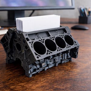 May include: A black, 3D-printed business card holder shaped like an engine block. The holder has five cylindrical openings and holds a stack of white business cards. The design is detailed and sits on a wooden desk.