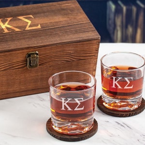 Può includere: Due bicchieri di whisky in vetro trasparente con le lettere "KS" incise sul davanti. I bicchieri sono appoggiati su sottobicchieri di legno e si trovano davanti a una scatola di legno con le stesse lettere incise sul coperchio.