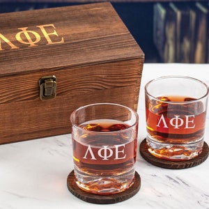 May include: Two clear glass whiskey tumblers with a wooden coaster each. The glasses are engraved with the Greek letters "ΛΦΕ". A wooden box with the same engraving is in the background.