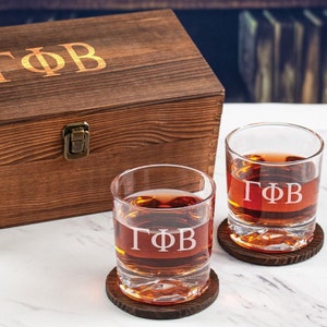May include: Two clear glass whiskey tumblers with the letters "ΓΦΒ" etched on the front. The glasses are sitting on wooden coasters and are in front of a wooden box with the same letters engraved on the lid.