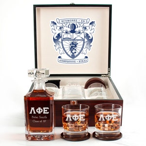 May include: A wooden box set containing a decanter and four glasses. The decanter and glasses are engraved with the Greek letters "ΛΦΕ" and the text "Peter Smith Class of '87". The box is lined with brown velvet and features a crest on the lid.