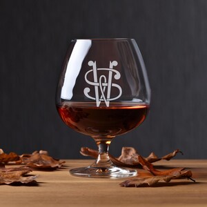 May include: A clear glass brandy snifter with a stem and a wide bowl. The glass is filled with a dark amber liquid. The glass has a monogram etched into the side, featuring the letters "S" and "W" intertwined.