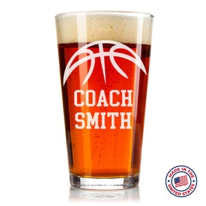 May include: A clear glass beer mug with a basketball design and the text "COACH SMITH" etched on the side. The mug is filled with a dark amber colored beer. The mug is made in the United States.