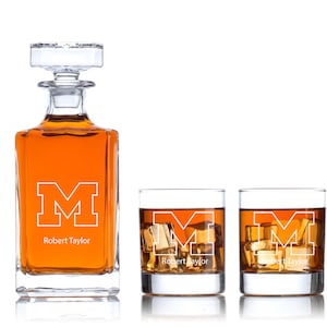 May include: A set of three clear glass decanter and glasses with a white outline of the letter "M" and the name "Robert Taylor" on each piece. The decanter is filled with amber liquid.