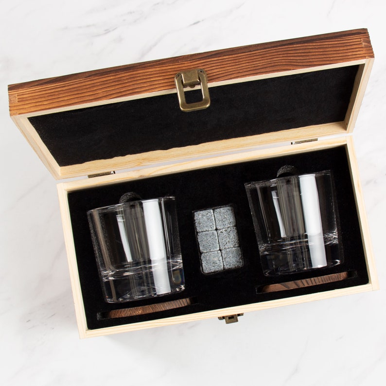 May include: A wooden box with a black interior contains two clear glass tumblers and a set of grey stone ice cubes. The box has a brass latch.
