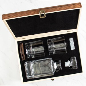 May include: A wooden box with a black interior, containing a clear glass decanter, two clear glass tumblers, and four gray stone ice cubes. The box is open and the items are visible.