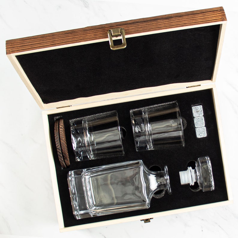 May include: A wooden box with a black interior, containing a clear glass decanter, two clear glass tumblers, and four gray stone ice cubes. The box is open and the items are visible.
