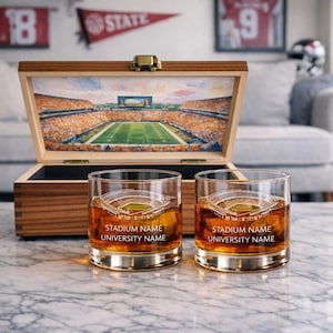 May include: Two whiskey glasses with a stadium design and the text "STADIUM NAME UNIVERSITY NAME" sit on a marble surface. A wooden box with a stadium painting inside is behind the glasses. The box is open, revealing the interior.