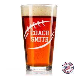 May include: A clear glass beer stein with a white football design and the text "COACH SMITH". The stein is filled with a dark amber beer and has a white foam head. The stein is made in the USA.