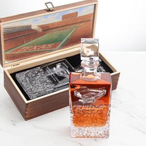 May include: A wooden box with a hinged lid, featuring a painting of a stadium and a football field. Inside, a glass decanter with a square stopper and a matching glass coaster are visible. The decanter has an etched stadium design and the text "Darrell K Royal Stadium".