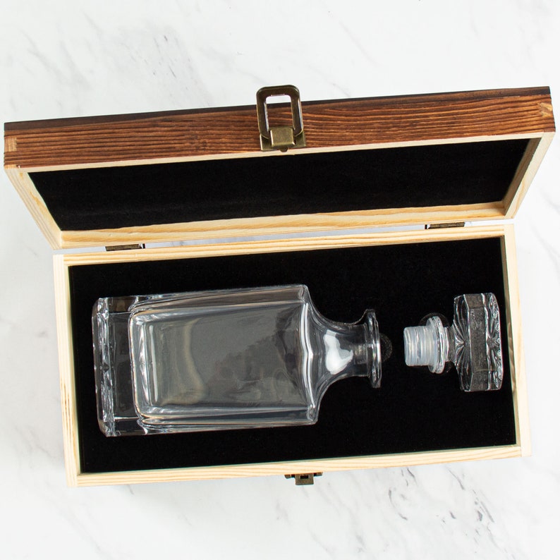 May include: A wooden box with a hinged lid, open to reveal a clear glass decanter with a stopper. The decanter is sitting on a black velvet lining inside the box.