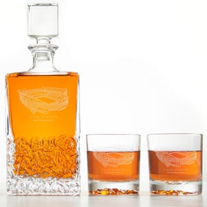 May include: A clear glass decanter with a square stopper and two matching lowball glasses. Each piece features an etched stadium design and the text "TIGER STADIUM". The decanter is filled with amber liquid.