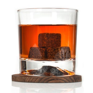 May include: A glass of amber-colored whiskey with three dark brown whiskey stones in the glass. The glass is sitting on a wooden coaster.