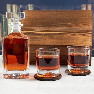 May include: A set of two clear glass whiskey tumblers with brown coasters and a clear glass decanter filled with amber liquid. The decanter has a stopper and sits on a wooden surface.