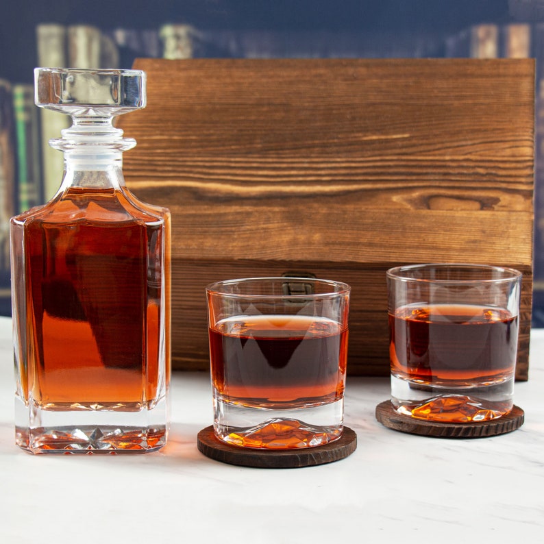 May include: A set of two clear glass whiskey tumblers with brown coasters and a clear glass decanter filled with amber liquid. The decanter has a stopper and sits on a wooden surface.