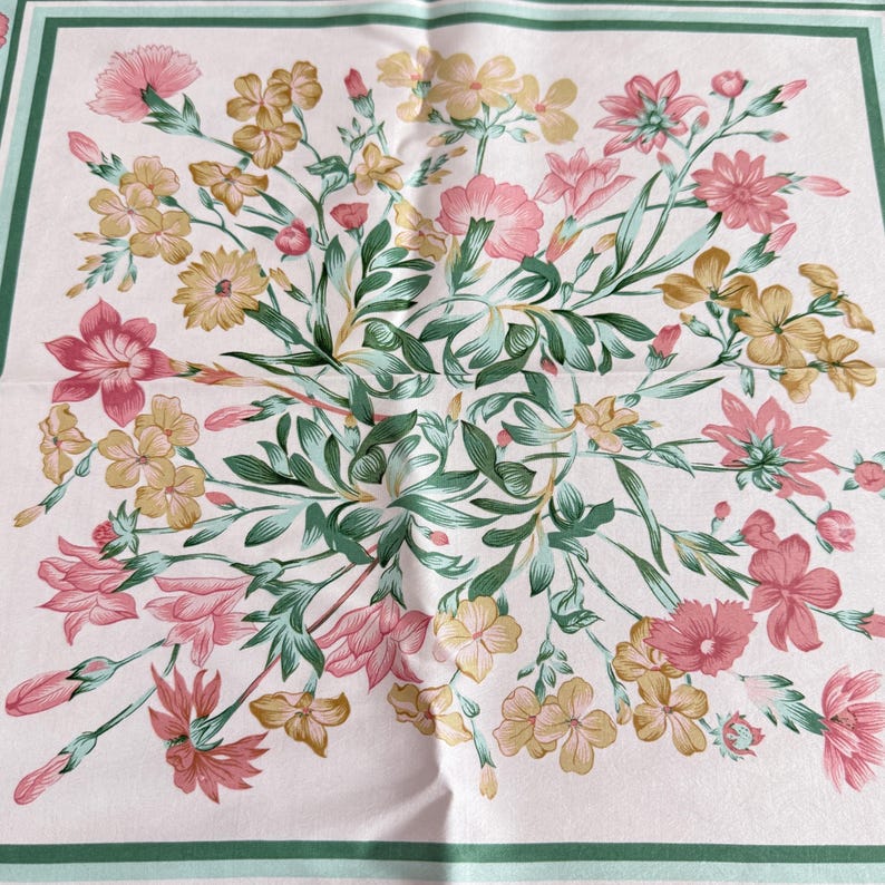 May include: A square scarf featuring a pale pink background with a central floral arrangement of pink and yellow flowers, green leaves, and stems. The design is framed by a double border in shades of dark and light green, creating a structured look.