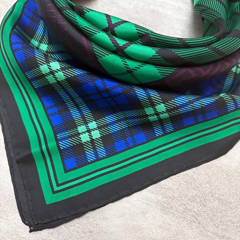May include: A square scarf with a plaid pattern in shades of green, blue, and black. The scarf has a black border with a green stripe. The plaid design features intersecting lines and squares.