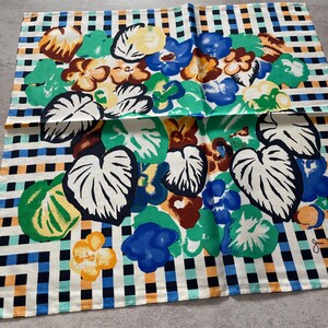 Op de afbeelding: Een witte katoenen theedoek met een geruit patroon in zwart, blauw en oranje. Het midden van de theedoek heeft een kleurrijk bloemenontwerp met groene bladeren, blauwe, oranje en bruine bloemen.