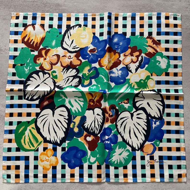Op de afbeelding: Een witte katoenen stof met een zwart-oranje ruitpatroon. De stof heeft een kleurrijk bloemenontwerp met groene bladeren, blauwe bloemen en bruine accenten. De stof is gesigneerd met "Jan Thompson".