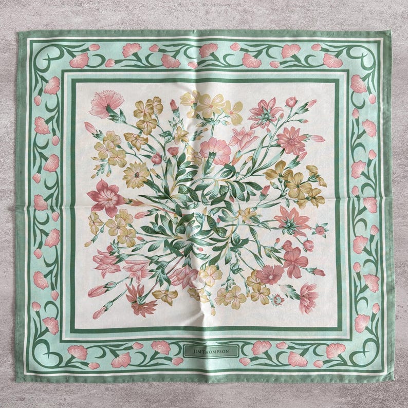May include: A square scarf featuring a central bouquet of pink and yellow flowers with green foliage on a white background. The border is a light green frame decorated with a repeating pattern of small pink flower buds and the text JIM THOMPSON.