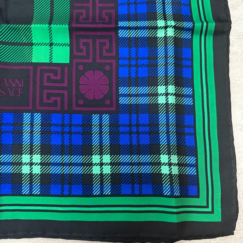 May include: A black silk scarf with a plaid pattern in shades of blue, green, and black. The scarf features a square design with geometric shapes in purple and the text "GIANNI VERSACE". The border is green.