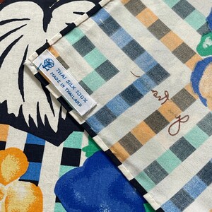 Op de afbeelding: Een close-up van een witte stof met een geruit patroon in tinten blauw, groen en bruin. De stof heeft een label met de tekst "Thai Silk 100% Made in Thailand".