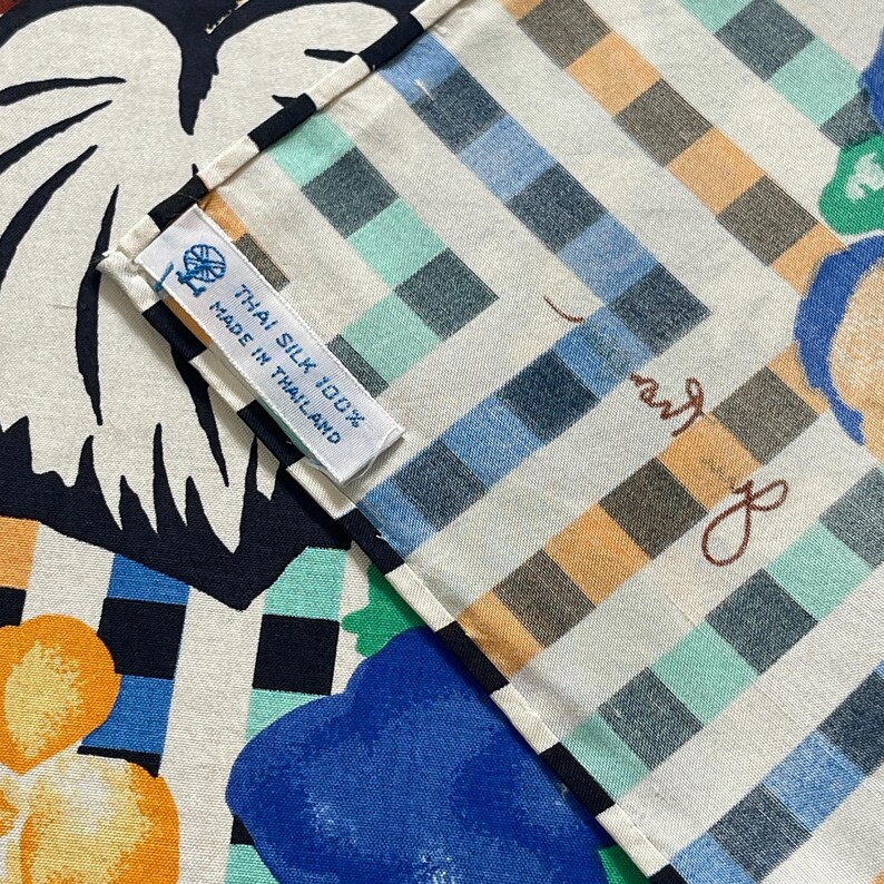 Op de afbeelding: Een close-up van een witte stof met een geruit patroon in tinten blauw, groen en bruin. De stof heeft een label met de tekst "Thai Silk 100% Made in Thailand".
