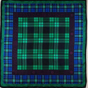 May include: A square scarf featuring a tartan pattern in green, blue, and black. The scarf has a border with geometric designs and a dark green edge. The central design is a large plaid.