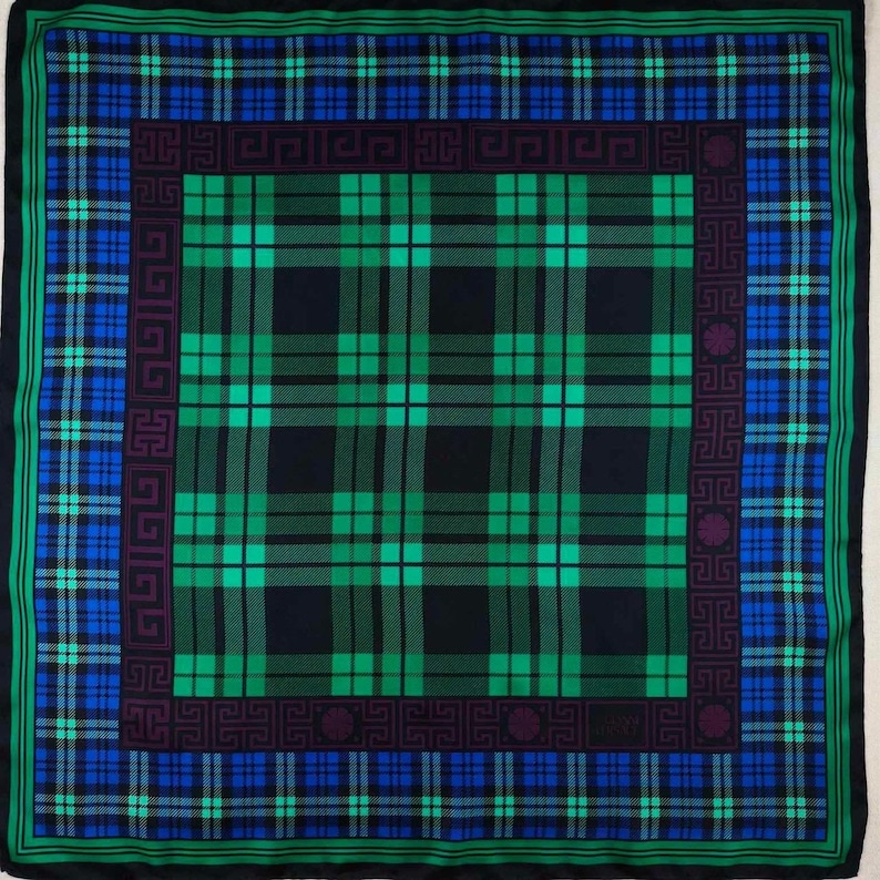 May include: A square scarf featuring a tartan pattern in green, blue, and black. The scarf has a border with geometric designs and a dark green edge. The central design is a large plaid.