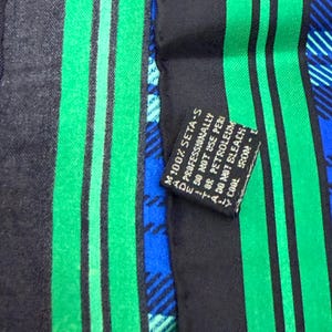 May include: A close-up of a silk scarf with a black background and vertical green stripes. The scarf also features blue plaid patterns. A small black tag reads "100% SETA-S" and care instructions.