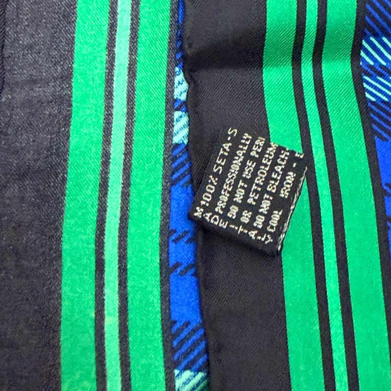 May include: A close-up of a silk scarf with a black background and vertical green stripes. The scarf also features blue plaid patterns. A small black tag reads "100% SETA-S" and care instructions.