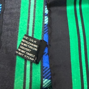 May include: A silk scarf with a pattern of black and green vertical stripes. A small black tag with white text is attached. The scarf also features blue and green plaid sections.