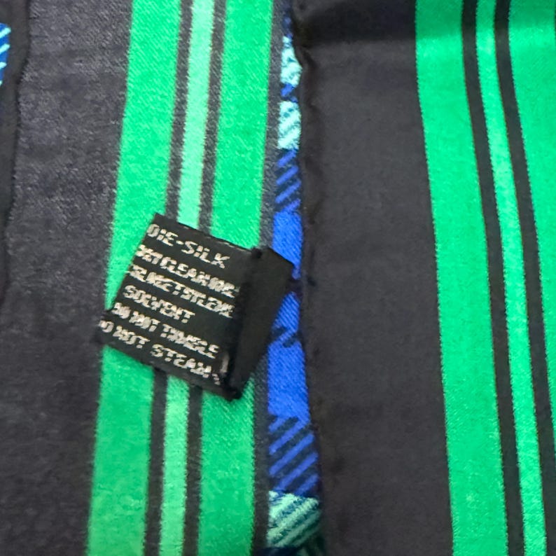 May include: A silk scarf with a pattern of black and green vertical stripes. A small black tag with white text is attached. The scarf also features blue and green plaid sections.