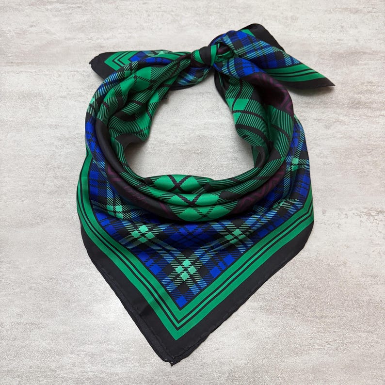 May include: A square scarf featuring a green, blue, and black tartan pattern. The scarf is knotted at the top, displaying the vibrant colours and design. The edges are trimmed with a solid black border, adding a defined finish to the accessory.