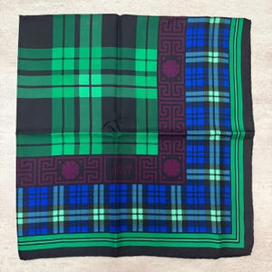 May include: A square silk scarf with a plaid pattern in black, green, and blue. The scarf features a border with a geometric design and the text "GIANNI VERSACE".