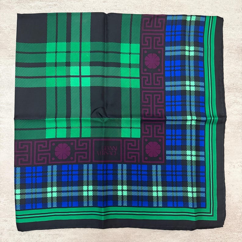 May include: A square silk scarf with a plaid pattern in black, green, and blue. The scarf features a border with a geometric design and the text "GIANNI VERSACE".