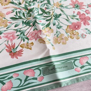 May include: A white fabric scarf featuring a floral pattern with pink, yellow, and green botanical illustrations. The design includes a border with a light green stripe and the text JIM THOMPSON printed in a dark green rectangular frame.
