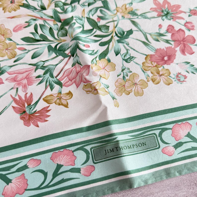 May include: A white fabric scarf featuring a floral pattern with pink, yellow, and green botanical illustrations. The design includes a border with a light green stripe and the text JIM THOMPSON printed in a dark green rectangular frame.