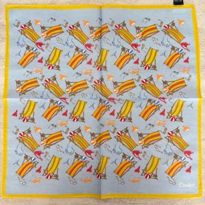 May include: A light blue handkerchief with a repeating pattern of beach chairs, beach balls, sunglasses, and bikinis. The handkerchief has a yellow border and the Drake's logo in the bottom right corner.