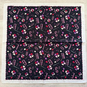 May include: A black and white handkerchief featuring a repeating pattern of hearts, the letter "G", and the word "LOVE". The border is white with a scalloped edge. The text "LULU GUINNESS" is also printed on the handkerchief.