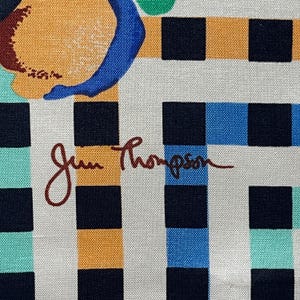 Op de afbeelding: Een witte stof met een zwart, oranje, blauw en groen geruit patroon. De stof is gesigneerd met "Jim Thompson" in bruine inkt.