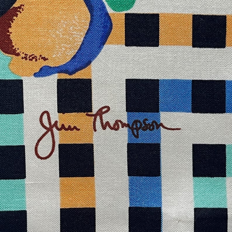 Op de afbeelding: Een witte stof met een zwart, oranje, blauw en groen geruit patroon. De stof is gesigneerd met "Jim Thompson" in bruine inkt.