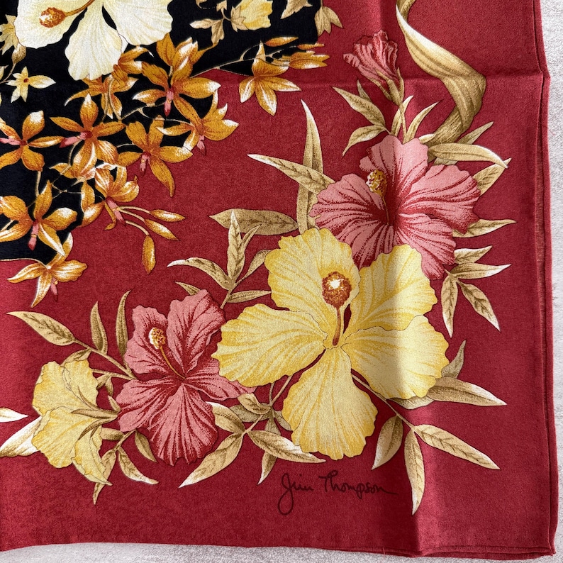 K&ouml;nnte beinhalten: Quadratisches rotes Seidentuch mit floralem Design. Der Schal zeigt gelbe, rosa und wei&szlig;e Hibiskusbl&uuml;ten mit einem schwarzen Abschnitt mit kleineren orangefarbenen Bl&uuml;ten. Die Signatur Jim Thompson befindet sich unten rechts.